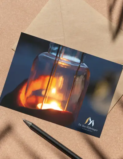Umschlag mit Karte einer brennenden Glaslampe in Hand, Logo und ‚Dr. Ina Mähringer Coaching‘ auf braunem Holztisch mit Stift