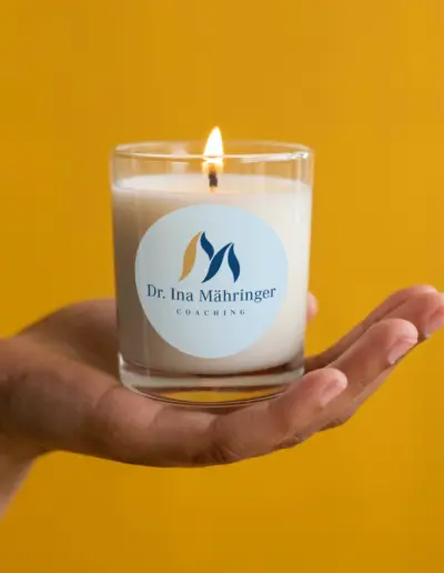Hand hält brennende Duftkerze mit weißem Etikett ‚Dr. Inna Mähhringerer Coaching‘ und Flammen-Symbol auf gelbem Hintergrund