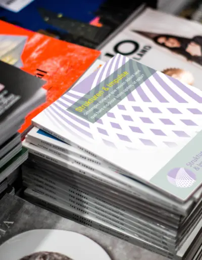 Stapel von ‚Strukturen & Impulse‘-Büchern mit violettem Design und Logo, umgeben von Magazinen wie ‚Gather Journal‘ und ‚The Senses‘ auf Holztisch