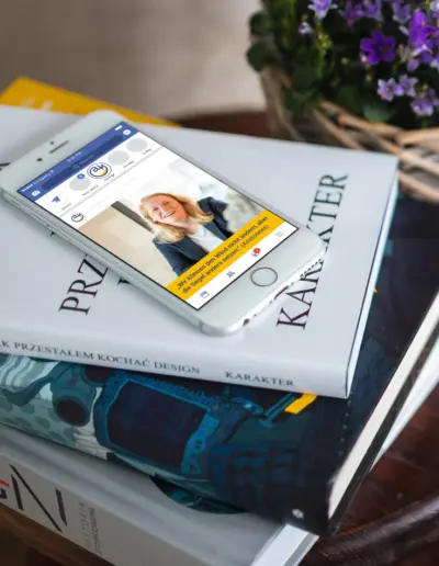 iPhone zeigt Website einer lächelnden Frau in Anzug mit gelbem Call-to-Action, neben Büchern wie ‚Jak Przestałem Kochać Design‘ und lila Blumen auf Tisch