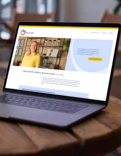 Anja Kurath: Laptop zeigt Website mit gelbem Logo, Foto einer Therapeutin in gelber Bluse, blauer Call-to-Action-Box und Zitat auf hellem Hintergrund auf Holztisch